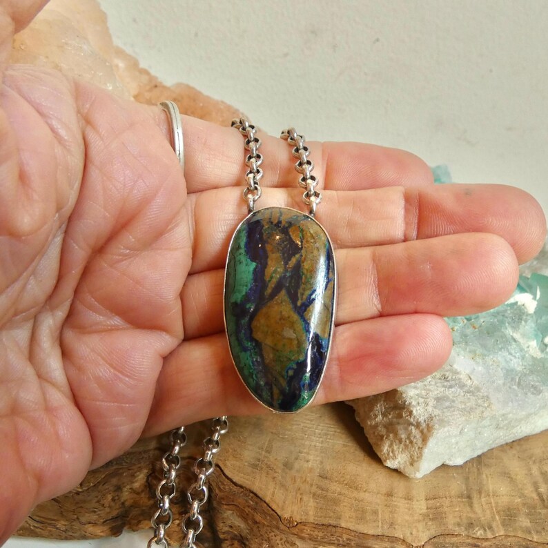 May include: A pendant necklace featuring an oval-shaped azurite-malachite gemstone with swirling patterns of deep blue, green, and brown. The stone is set in a smooth silver bezel and hangs from a thick, round silver link chain.