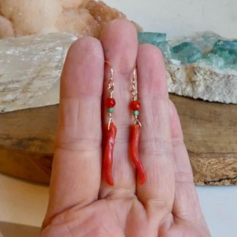 May include: A pair of dangle earrings featuring long, curved, red coral-colored pendants suspended from gold-toned metal hooks. Each earring includes a small, faceted red bead and a tiny turquoise-colored accent bead above the main pendant.