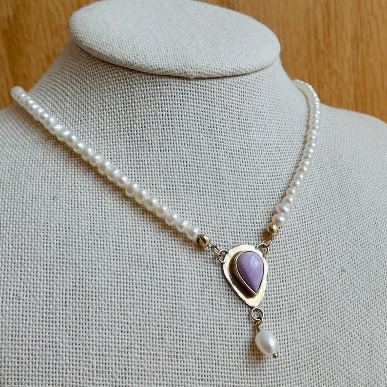 Puede incluir: Un delicado collar de perlas con un colgante dorado con una piedra en forma de l&aacute;grima de color p&uacute;rpura claro. Una sola perla cuelga de la parte inferior del colgante. El collar se exhibe en un maniqu&iacute;.
