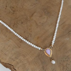 Puede incluir: Un delicado collar con una hebra de peque&ntilde;as perlas blancas. El colgante tiene un marco dorado con una piedra en forma de l&aacute;grima de color lila claro y una perla blanca colgante. El collar se exhibe sobre una superficie de madera.