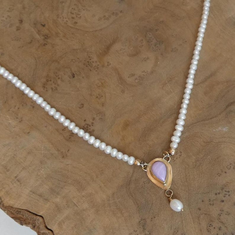 Puede incluir: Un delicado collar con una hebra de peque&ntilde;as perlas blancas. El colgante tiene un marco dorado con una piedra en forma de l&aacute;grima de color lila claro y una perla blanca colgante. El collar se exhibe sobre una superficie de madera.
