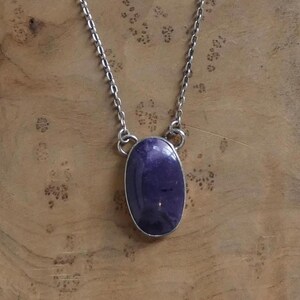 May include: A silver-toned necklace featuring a large, oval-shaped, deep purple gemstone pendant. The pendant is set in a silver bezel and hangs from a delicate chain. The necklace is displayed on a wooden surface.