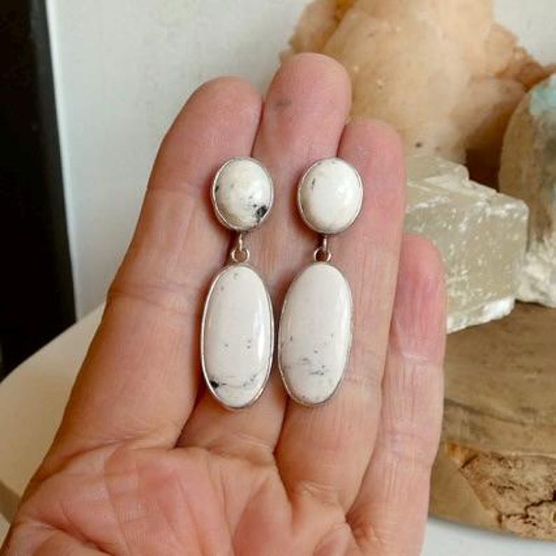 May include: A pair of drop earrings featuring two white oval stones with black inclusions set in silver-toned metal. Each earring consists of a smaller round stone at the top connected to a larger elongated oval stone hanging below.