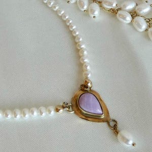 Puede incluir: Un delicado collar con una cadena de peque&ntilde;as perlas blancas. El punto focal es un colgante dorado con una piedra en forma de l&aacute;grima de color lavanda. Una sola perla cuelga del colgante. El collar est&aacute; sobre un fondo blanco.