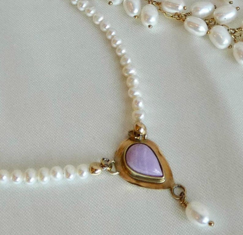 Puede incluir: Un delicado collar con una cadena de peque&ntilde;as perlas blancas. El punto focal es un colgante dorado con una piedra en forma de l&aacute;grima de color lavanda. Una sola perla cuelga del colgante. El collar est&aacute; sobre un fondo blanco.