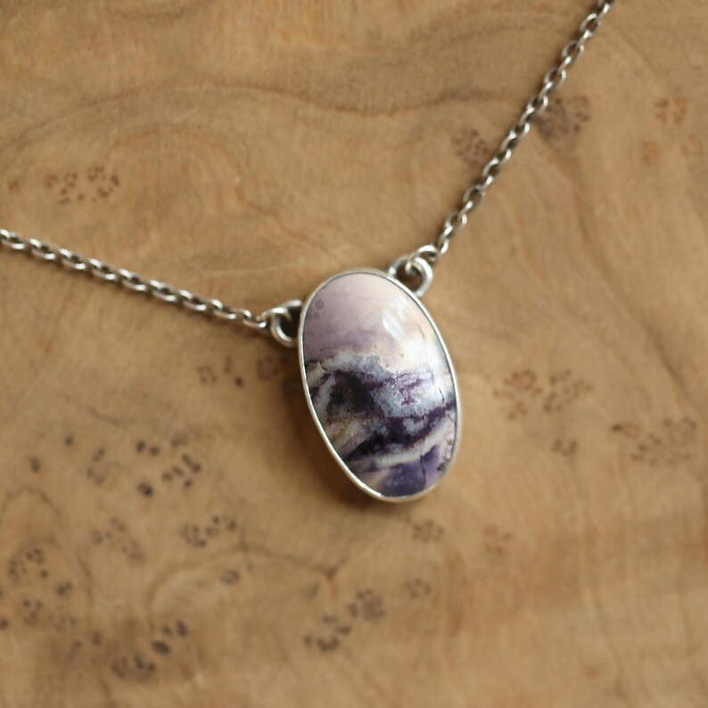 May include: A silver chain necklace featuring an oval pendant. The pendant has a light purple and white stone with dark purple and black veining. The necklace is displayed on a wooden surface.