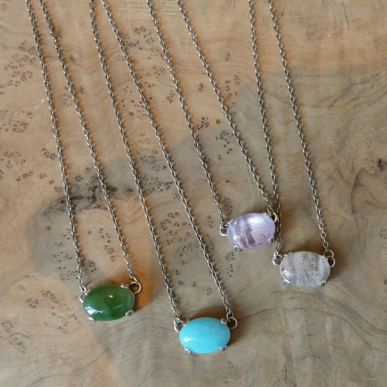 K&ouml;nnte beinhalten: Vier Halsketten mit d&uuml;nnen Metallketten und ovalen Cabochon-Steinen in Gr&uuml;n, Hellblau, Hellviolett und klarem Wei&szlig;. Jeder Anh&auml;nger wird von einer kleinen Metallfassung gehalten, die auf beiden Seiten mit der Kette verbunden ist.