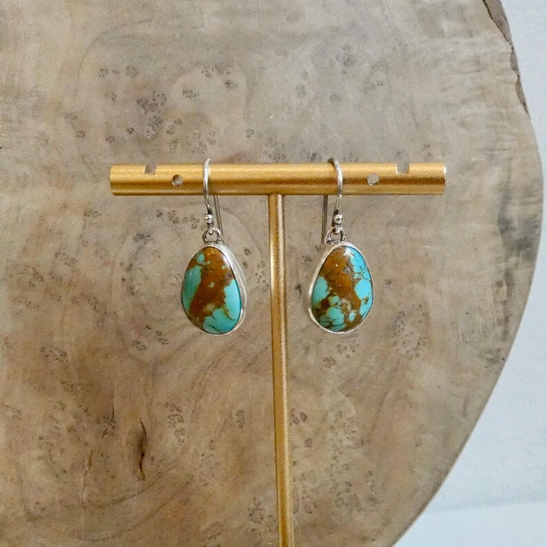Puede incluir: Un par de pendientes en forma de l&aacute;grima con piedras de turquesa pulidas con patrones de matriz marr&oacute;n, engastados en marcos de metal plateado con ganchos, suspendidos de una barra de exhibici&oacute;n horizontal de metal dorado.