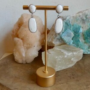 May include: A pair of drop earrings featuring two white oval stones each, set in silver-toned metal. The top stone is smaller and circular, while the bottom stone is larger and elongated. The earrings are displayed on a gold-toned metal stand.