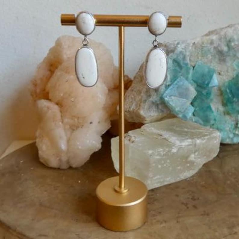 May include: A pair of drop earrings featuring two white oval stones each, set in silver-toned metal. The top stone is smaller and circular, while the bottom stone is larger and elongated. The earrings are displayed on a gold-toned metal stand.