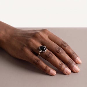 May include: A silver ring featuring a smooth, oval-shaped black stone set in a twisted metal frame. The band is thin and metallic, showcasing a polished finish that encircles the finger, highlighting the contrast between the dark stone and silver.