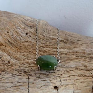 K&ouml;nnte beinhalten: Eine Halskette mit einem glatten, ovalen, dunkelgr&uuml;nen Edelstein in einer silberfarbenen Metallfassung mit Krappen. Der Anh&auml;nger ist an einer zierlichen silberfarbenen Metallkette mit kleinen, gleichm&auml;&szlig;igen, runden Gliedern befestigt.