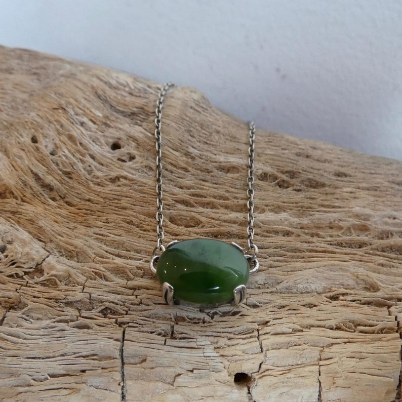 K&ouml;nnte beinhalten: Eine Halskette mit einem glatten, ovalen, dunkelgr&uuml;nen Edelstein in einer silberfarbenen Metallfassung mit Krappen. Der Anh&auml;nger ist an einer zierlichen silberfarbenen Metallkette mit kleinen, gleichm&auml;&szlig;igen, runden Gliedern befestigt.