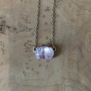 K&ouml;nnte beinhalten: Eine Halskette mit einem polierten, ovalen, hellvioletten Edelstein in einer silberfarbenen Metallfassung mit Krappen. Der Anh&auml;nger h&auml;ngt an einer zarten, d&uuml;nnen silberfarbenen Metallkette mit kleinen, gleichm&auml;&szlig;igen ovalen Gliedern.