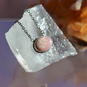 K&ouml;nnte beinhalten: Eine Halskette mit einem ovalen, hellrosa Stein, der in eine glatte, silberfarbene Metallfassung eingelassen ist. Der Anh&auml;nger ist an einer zarten, silberfarbenen Metallkette mit kleinen, gleichm&auml;&szlig;igen runden Gliedern befestigt.