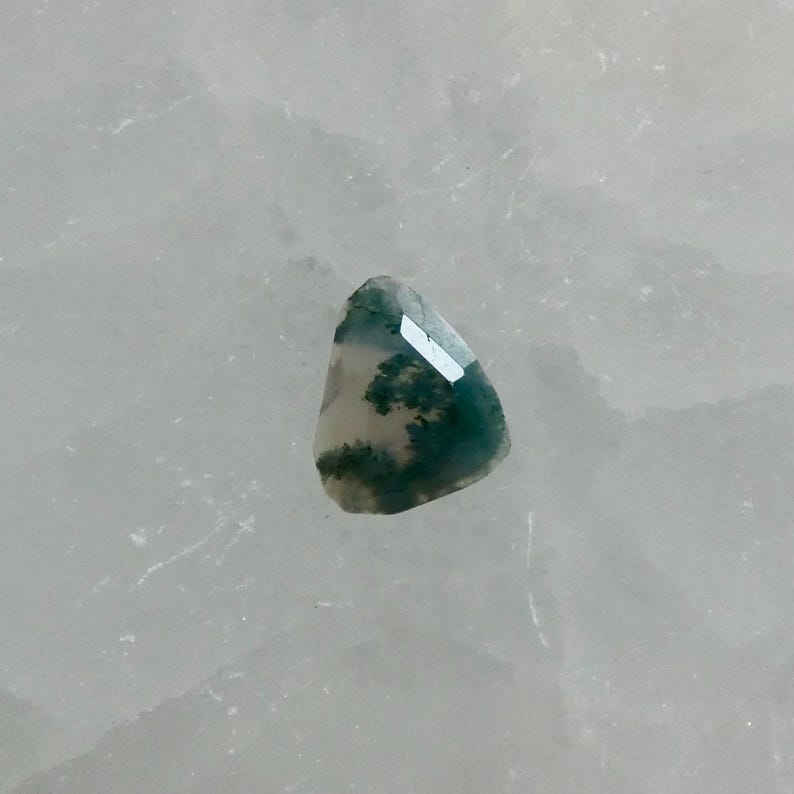 May include: A faceted moss agate gemstone with a translucent white base and deep green, tree-like dendritic inclusions. The stone has a triangular shape with rounded edges and a polished surface that reflects light across its multiple flat facets.