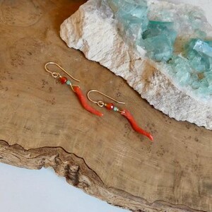 May include: A pair of gold-tone hook earrings featuring a curved, slender red coral-colored element, a small round orange bead, and a tiny turquoise-colored accent bead. The earrings are suspended from gold-colored wire hooks.