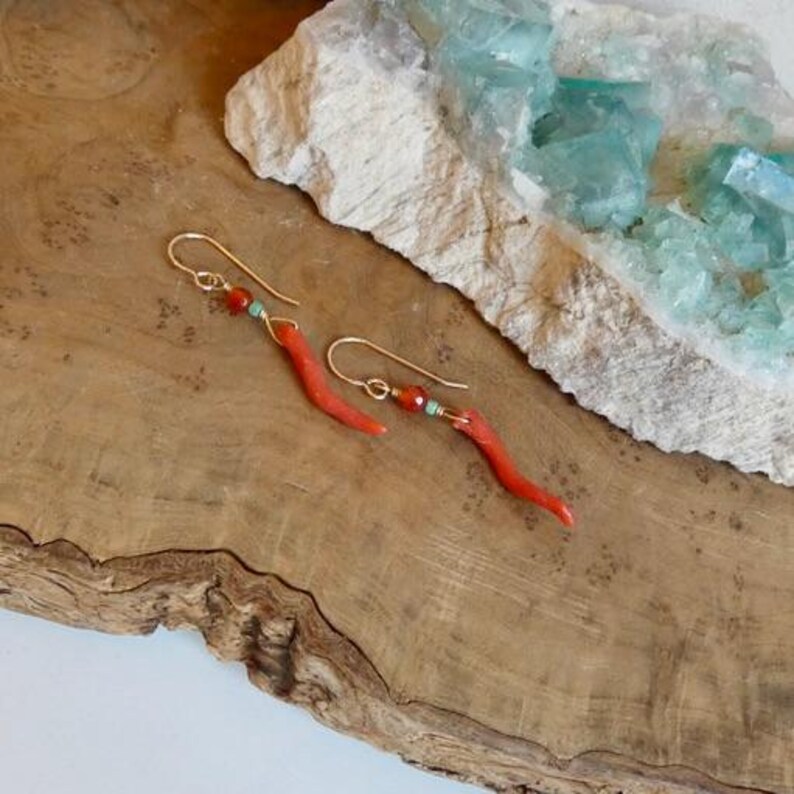 May include: A pair of gold-tone hook earrings featuring a curved, slender red coral-colored element, a small round orange bead, and a tiny turquoise-colored accent bead. The earrings are suspended from gold-colored wire hooks.