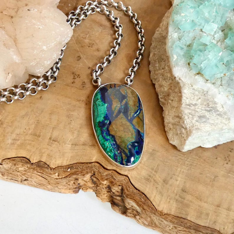 May include: A silver pendant necklace featuring an oval-shaped azurite and malachite gemstone with deep blue and vibrant green patterns. The stone is set in a smooth silver bezel and hangs from a silver rolo chain with round interlocking links.