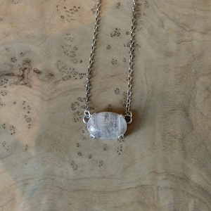 K&ouml;nnte beinhalten: Eine silberfarbene Metallkettenhalskette mit einem einzelnen, ovalen, durchscheinenden wei&szlig;en Steinanh&auml;nger. Der Stein wird von vier kleinen Metallkrallen gehalten, und die Kette ist an beiden Seiten der Fassung mit dem Anh&auml;nger verbunden.