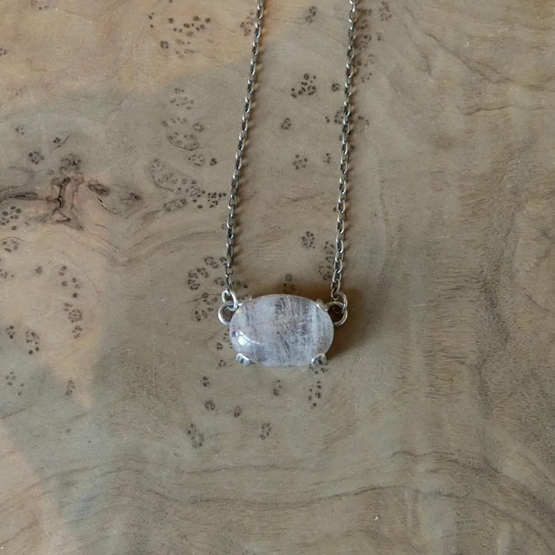 K&ouml;nnte beinhalten: Eine silberfarbene Metallkettenhalskette mit einem einzelnen, ovalen, durchscheinenden wei&szlig;en Steinanh&auml;nger. Der Stein wird von vier kleinen Metallkrallen gehalten, und die Kette ist an beiden Seiten der Fassung mit dem Anh&auml;nger verbunden.