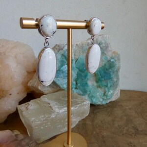 May include: A pair of drop earrings featuring smooth, oval-shaped white stones with subtle dark inclusions, set in polished silver-toned metal. Each earring consists of a smaller circular top stone and a larger oval bottom stone connected by a metal link.