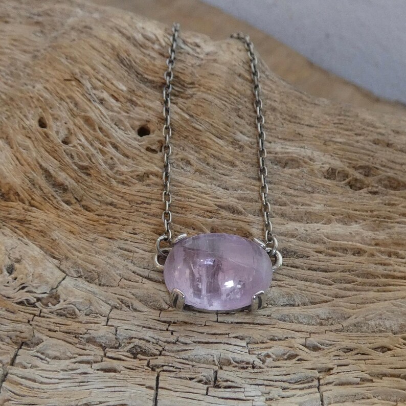 K&ouml;nnte beinhalten: Eine Halskette mit einem glatten, ovalen, durchscheinenden hellvioletten Edelstein in einer silberfarbenen Metallfassung. Der Anh&auml;nger ist an einer zarten, d&uuml;nnen silberfarbenen Metallkette mit kleinen, gleichm&auml;&szlig;igen ovalen Gliedern befestigt.