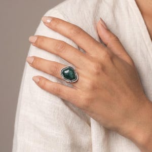 May include: A ring featuring a large, irregular, faceted dark green gemstone set in a polished silver band. The stone has a distinct geometric shape with multiple flat surfaces that catch the light, secured by a simple, smooth metal setting.