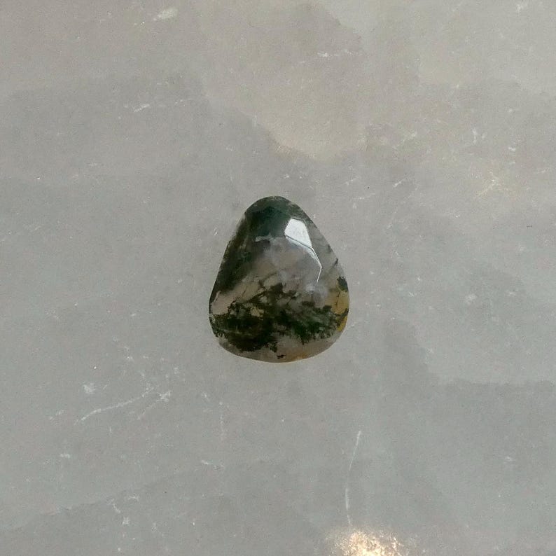 May include: A teardrop-shaped moss agate gemstone with a faceted surface. The stone features a translucent, milky white base with distinct dark green, fern-like inclusions spreading throughout the interior, creating a natural, organic pattern.