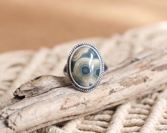 Può includere: Un anello in argento con una pietra cabochon di forma ovale caratterizzata da motivi circolari concentrici nei toni del grigio, beige e verde scuro. La pietra è incastonata in una lunetta liscia circondata da un bordo metallico intrecciato.
