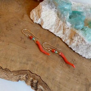 May include: A pair of gold-toned dangle earrings featuring a curved, branch-like red coral-colored element, a small round orange bead, and a tiny turquoise-colored accent bead suspended from a simple gold-toned wire hook for pierced ears.