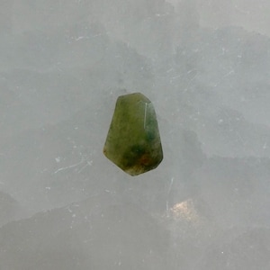 May include: A small, faceted gemstone with a translucent appearance, displaying a mix of olive green, light yellow, and subtle dark green inclusions throughout its irregular, multi-sided shape, set against a textured, pale white background.