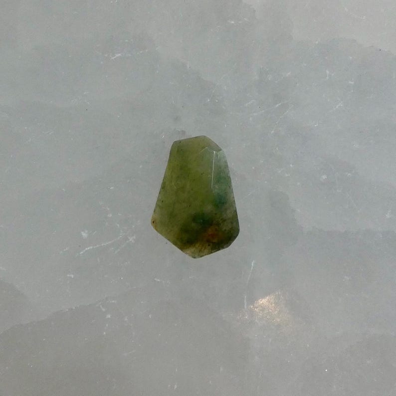 May include: A small, faceted gemstone with a translucent appearance, displaying a mix of olive green, light yellow, and subtle dark green inclusions throughout its irregular, multi-sided shape, set against a textured, pale white background.