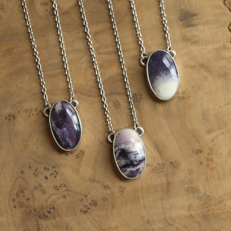 May include: Four silver chain necklaces with oval pendants. The pendants feature polished stones in shades of purple, lavender, and white. The necklaces are displayed on a wooden surface.