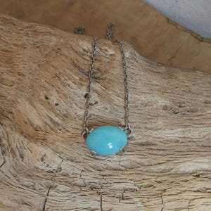 K&ouml;nnte beinhalten: Eine silberfarbene Metallkettenhalskette mit einem einzelnen, glatten, ovalen, t&uuml;rkisblauen Steinanh&auml;nger, der von vier kleinen Metallkrallen gehalten wird. Die Kette ist zart und besteht aus einfachen, gleichm&auml;&szlig;igen Gliedern.