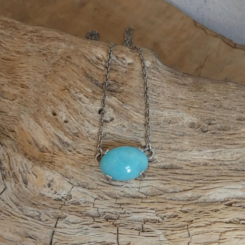 K&ouml;nnte beinhalten: Eine silberfarbene Metallkettenhalskette mit einem einzelnen, glatten, ovalen, t&uuml;rkisblauen Steinanh&auml;nger, der von vier kleinen Metallkrallen gehalten wird. Die Kette ist zart und besteht aus einfachen, gleichm&auml;&szlig;igen Gliedern.