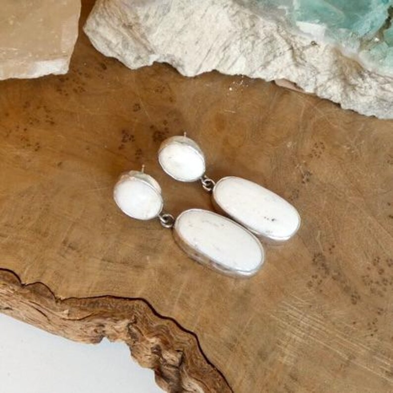 May include: A pair of drop earrings featuring two white, oval-shaped stones set in silver-toned metal. Each earring consists of a smaller circular stone at the top connected to a larger, elongated oval stone below, creating a dangling design.