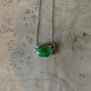 K&ouml;nnte beinhalten: Eine silberne Halskette mit einem einzelnen ovalen, glatten und polierten gr&uuml;nen Steinanh&auml;nger. Der Stein wird von einer silbernen Fassung mit vier Krappen gehalten, und die feine Metallkette ist an beiden Seiten der Steinfassung befestigt.
