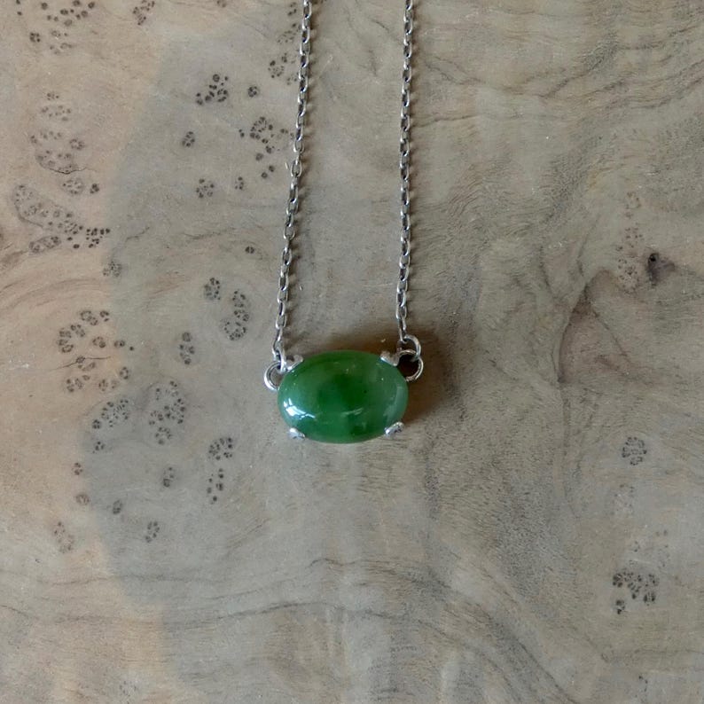 K&ouml;nnte beinhalten: Eine silberne Halskette mit einem einzelnen ovalen, glatten und polierten gr&uuml;nen Steinanh&auml;nger. Der Stein wird von einer silbernen Fassung mit vier Krappen gehalten, und die feine Metallkette ist an beiden Seiten der Steinfassung befestigt.
