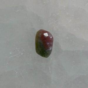 May include: A small, faceted gemstone with a smooth, rounded shape. The stone features a distinct color gradient, transitioning from a deep, earthy green at the base to a warm, reddish-pink hue at the top, with a small white inclusion visible.