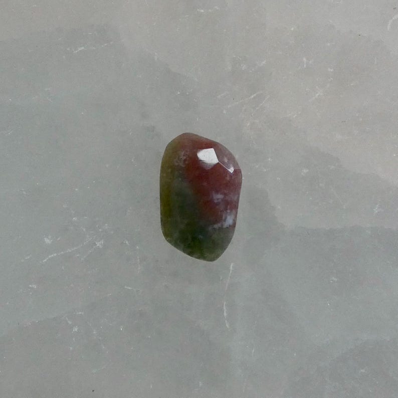 May include: A small, faceted gemstone with a smooth, rounded shape. The stone features a distinct color gradient, transitioning from a deep, earthy green at the base to a warm, reddish-pink hue at the top, with a small white inclusion visible.