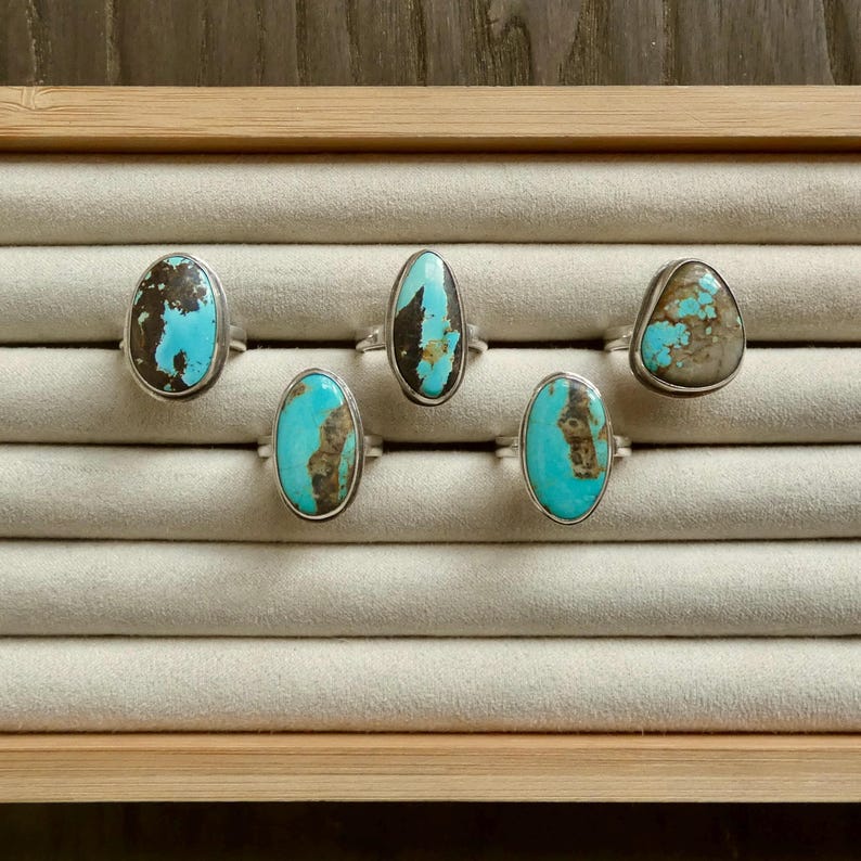 K&ouml;nnte beinhalten: F&uuml;nf Silberringe mit ovalen T&uuml;rkissteinen, die ausgepr&auml;gte braune Matrixmuster aufweisen. Jeder Ring zeigt einen einzigartigen, nat&uuml;rlich blau-gr&uuml;nen Edelstein in einer glatten Metallschiene, angeordnet in einer Reihe auf einem hellen Samt-Display.