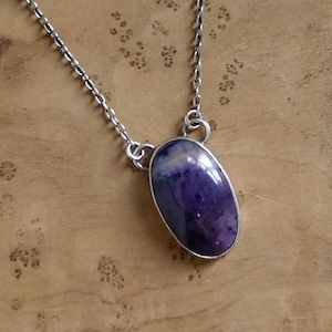 May include: A silver necklace featuring a large, oval-shaped purple gemstone pendant. The gemstone has a polished surface and is set in a silver bezel. The chain is a delicate silver link.