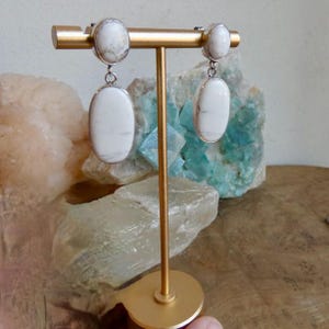 May include: A pair of drop earrings featuring smooth, oval-shaped white stones with subtle grey veining. Each earring consists of a smaller round stone at the top and a larger, elongated oval stone suspended below, set in polished silver-toned metal.
