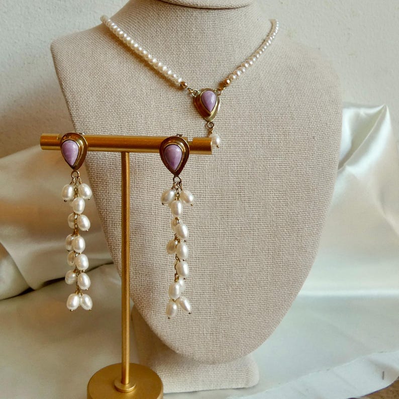 Puede incluir: Un conjunto de joyas vintage con un collar de perlas y pendientes a juego. El collar y los pendientes tienen un engaste de metal dorado con una piedra lavanda en forma de l&aacute;grima. Los pendientes tienen m&uacute;ltiples hilos de peque&ntilde;as perlas.