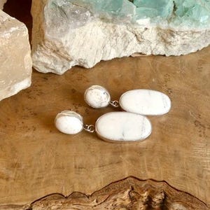 May include: A pair of drop earrings featuring white oval-shaped stones with grey veining, each set in a silver-toned metal frame. The earrings consist of a smaller round stone at the top connected to a larger, elongated oval stone at the bottom.