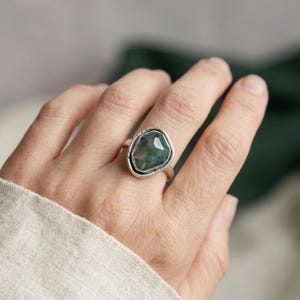 May include: A silver ring featuring a faceted, irregular-shaped moss agate gemstone with deep green inclusions. The stone is set in a smooth, polished silver bezel with a simple, slender band that wraps around the finger, showcasing a natural aesthetic.