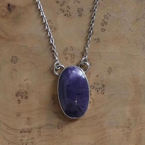 May include: A silver-toned necklace featuring a large, oval-shaped, deep purple gemstone pendant. The pendant is set in a silver bezel and hangs from a delicate chain. The gemstone has a polished surface, reflecting light. The necklace is displayed on a wooden surface.