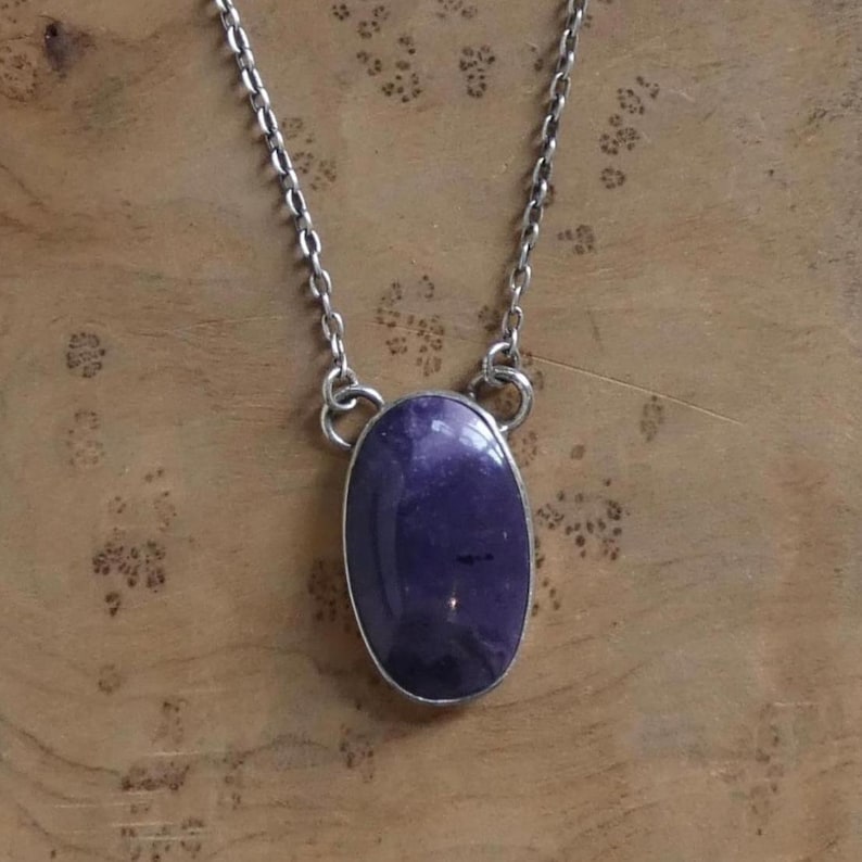 May include: A silver-toned necklace featuring a large, oval-shaped, deep purple gemstone pendant. The pendant is set in a silver bezel and hangs from a delicate chain. The gemstone has a polished surface, reflecting light. The necklace is displayed on a wooden surface.