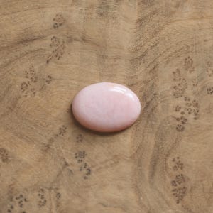 K&ouml;nnte beinhalten: Ein glatter, oval geformter rosa Edelstein mit polierter Oberfl&auml;che. Der Stein hat eine weiche, undurchsichtige hellrosa Farbe mit subtilen, wirbelnden Farbvariationen auf seiner abgerundeten Oberfl&auml;che, die das Licht gleichm&auml;&szlig;ig reflektiert.